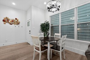 A dining area with a round table, white chairs, a chandelier, and a large window with blinds. - East End Stay | Near Daikin Park & Downtown (Houston)