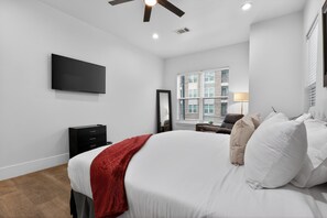 A modern bedroom with a large bed, a flat-screen TV, a dresser, and a window with a city view. - East End Stay | Near Daikin Park & Downtown (Houston)
