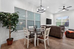 Ein modernes Wohnzimmer mit Essbereich, Deckenventilator und einem großen Fenster mit Jalousien.