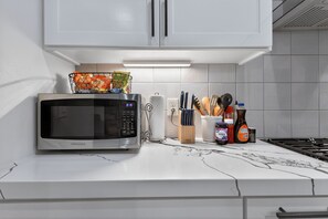 A kitchen with a microwave, a basket of snacks, a knife block, and condiments. - East End Stay | Near Daikin Park & Downtown (Houston)