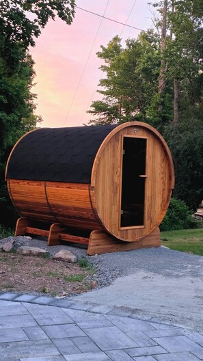 Een houten, vatvormig gebouw met een deur, gelegen op een verhard gebied met omliggend groen.