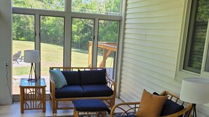 A sunlit room with a large window, a patterned rug, wicker furniture, and a table with a lamp.