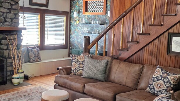 A cozy living room with a brown leather sofa, a stone fireplace, and a wooden staircase.