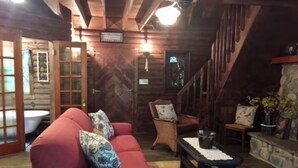 A log cabin interior with a red sofa, wicker chairs, a stone fireplace, and a wooden staircase.