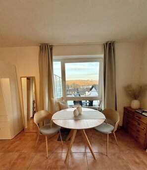 A dining area with a round table, two chairs, a window with curtains, and a view of buildings.
