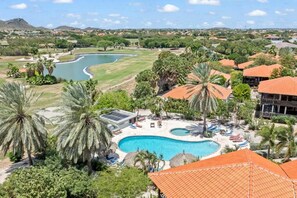 Een resort met een zwembad, palmbomen en een golfbaan op de achtergrond.