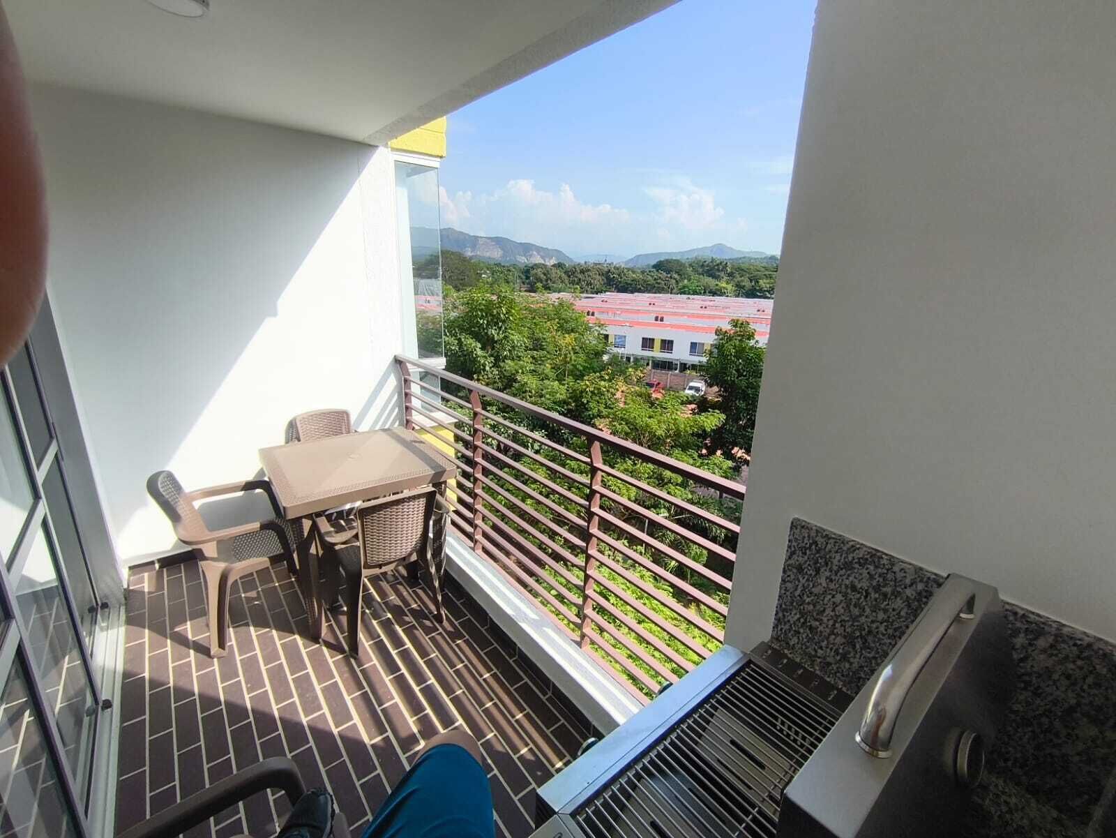 A balcony with a table and chairs, a grill, and a view of buildings and greenery.