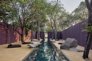 Ein ruhiger Poolbereich im Freien mit einem klaren Wasserlauf, umgeben von Bäumen und großen Steinen, mit violetten Wänden und sandigem Untergrund.
