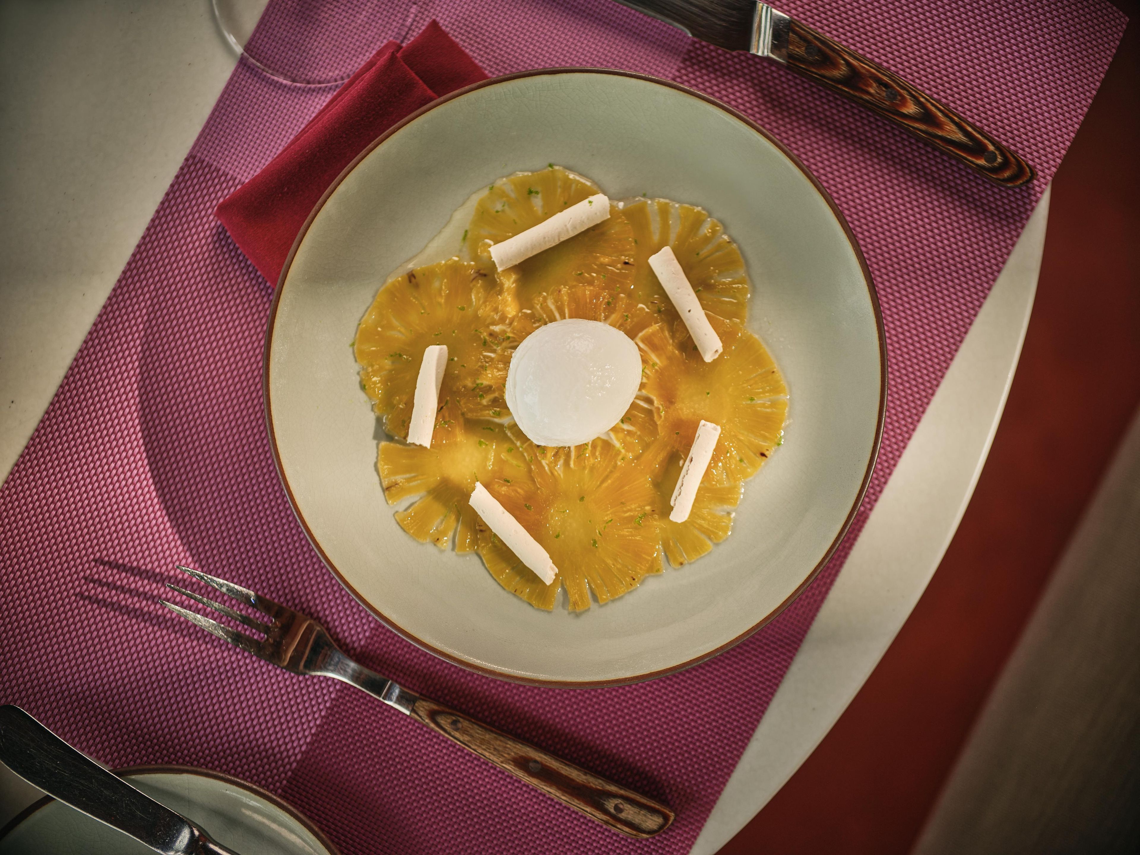 Ein Teller mit Ananasstückchen und einem weißen Objekt, vermutlich ein Dessert, auf einem Tisch mit einer roten Servette und einer Gabel.