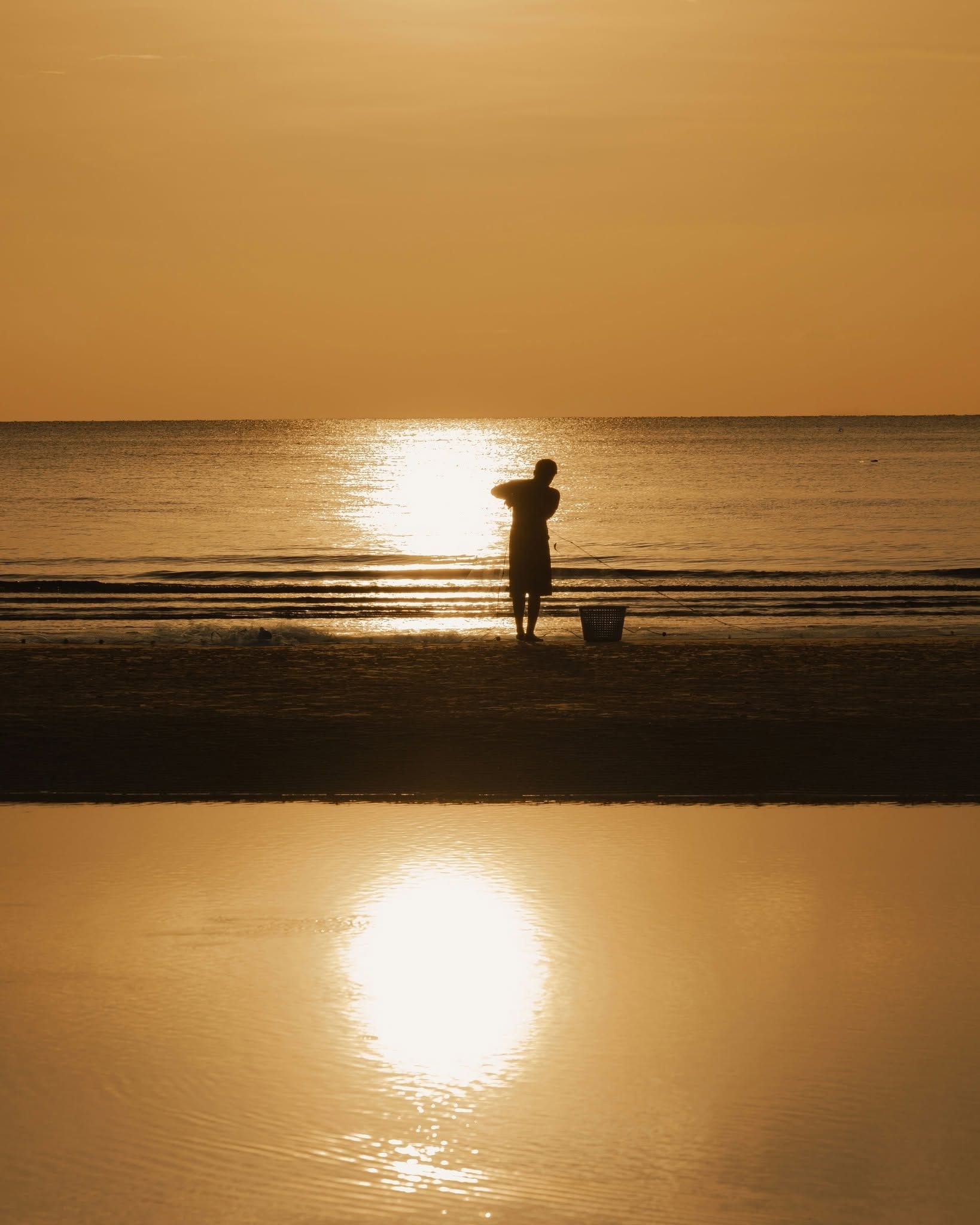 A beach scene with a person standing near the water's edge, a basket, and the sun reflecting on the water.