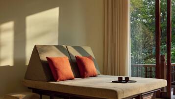 A room with a sofa bed, two pillows, a tray, and a view of greenery through large windows.
