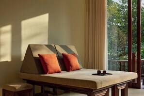 A room with a sofa bed, two pillows, a tray, and a view of greenery through large windows.