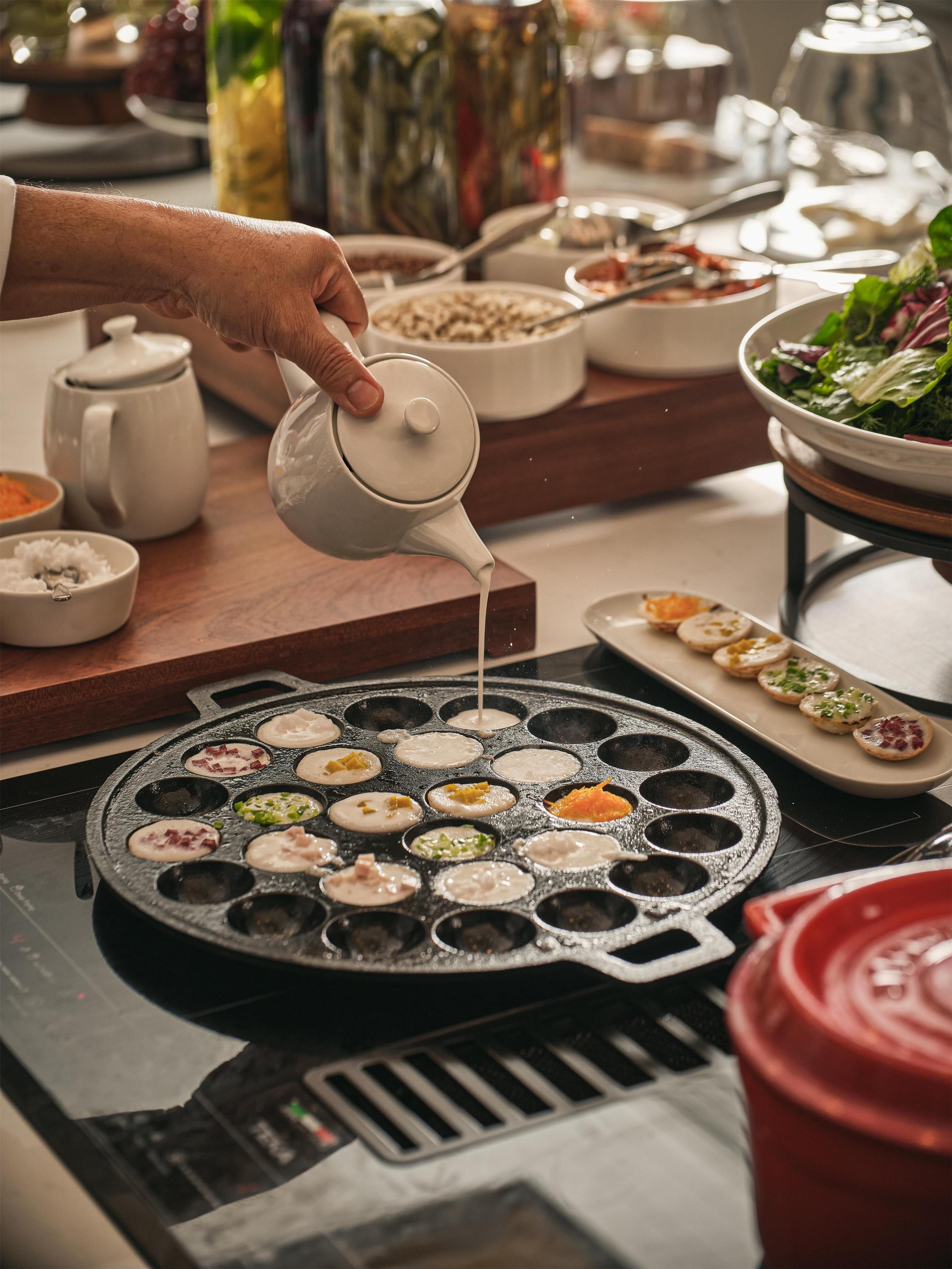 Une personne verse un liquide sur une surface de cuisson chauffée, sur laquelle se trouvent plusieurs moules ronds.