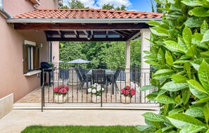 Eine überdachte Terrasse mit Tisch und Stühlen, umgeben von einem Metallzaun und Blumentöpfen.