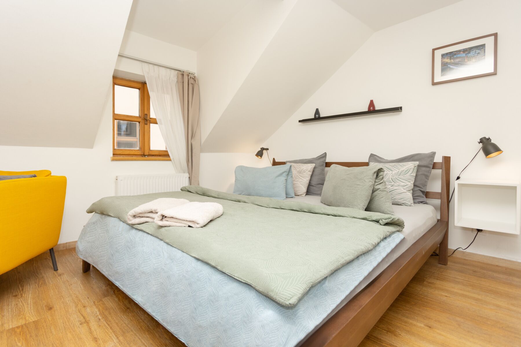 A bedroom with a bed, a window, a bedside table, a lamp, and a picture on the wall.