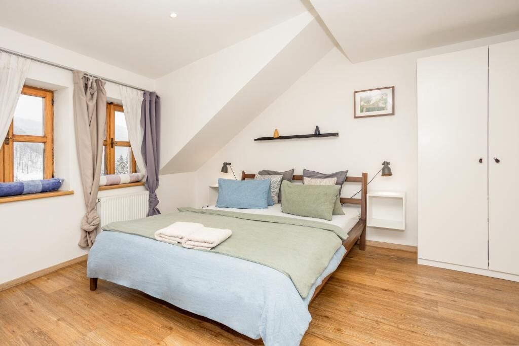 A bedroom with a bed, a window with curtains, a radiator, a shelf with a picture, and a wardrobe.