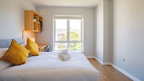 A bedroom with a bed, two pillows, a bedside table, a lamp, a shelf with a plant, and a window. - Prime Location Townhouse Galway (Galway)