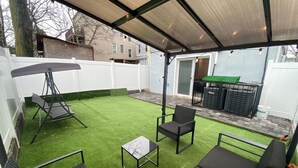 Eine Außenpatio mit Kunstgrasanlage, einem überdachten Sitzbereich und Blick auf ein Wohngebäude.