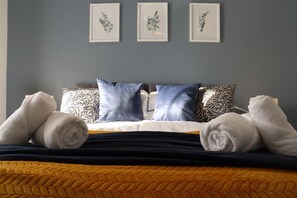 A bed with rolled towels, a patterned pillow, and framed botanical prints on the wall.