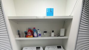A laundry room with two front-loading washers and dryers, a shelf with cleaning supplies, and a power outlet. - The Boyer House Beautiful Modern Farmhouse (Paducah)