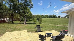 A covered deck with outdoor furniture, a grill, and a view of a grassy field and trees. - The Boyer House Beautiful Modern Farmhouse (Paducah)