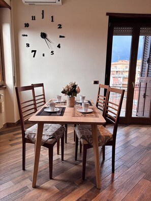 Een eetruimte met een houten tafel, vier stoelen, een wandklok en uitzicht op gebouwen door een glazen deur.