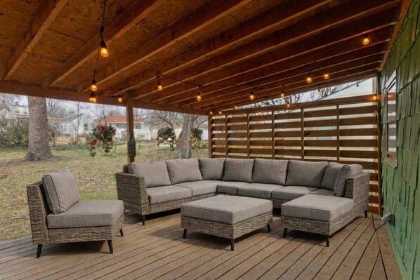 Un espace extérieur aménagé, abrité et doté d’un canapé d’angle et de deux fauteuils, d’un plancher en bois et de guirlandes lumineuses.