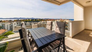 Un balcón con mesa y sillas, con vista a un paisaje urbano con edificios y una vista distante del mar.