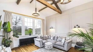 A living room with a sectional sofa, a round ottoman, a wooden floor, a radiator, and a large window with curtains. - Farm House - Sleeps 16 - Hot Tub - Pet Friendly (Skipton)