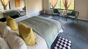 Ein modernes Schlafzimmer mit einem großen Bett, einem kleinen Tisch, zwei Stühlen und Blick ins Freie durch große Fenster.