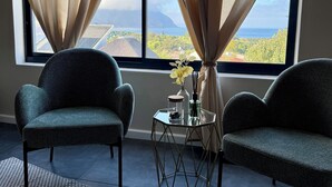 Ein Zimmer mit zwei grünen Sitzmöglichkeiten, einem kleinen Glastisch mit einer Vase und einem Glas, und Blick auf eine Landschaft durch große Fenster mit beigen Vorhängen.
