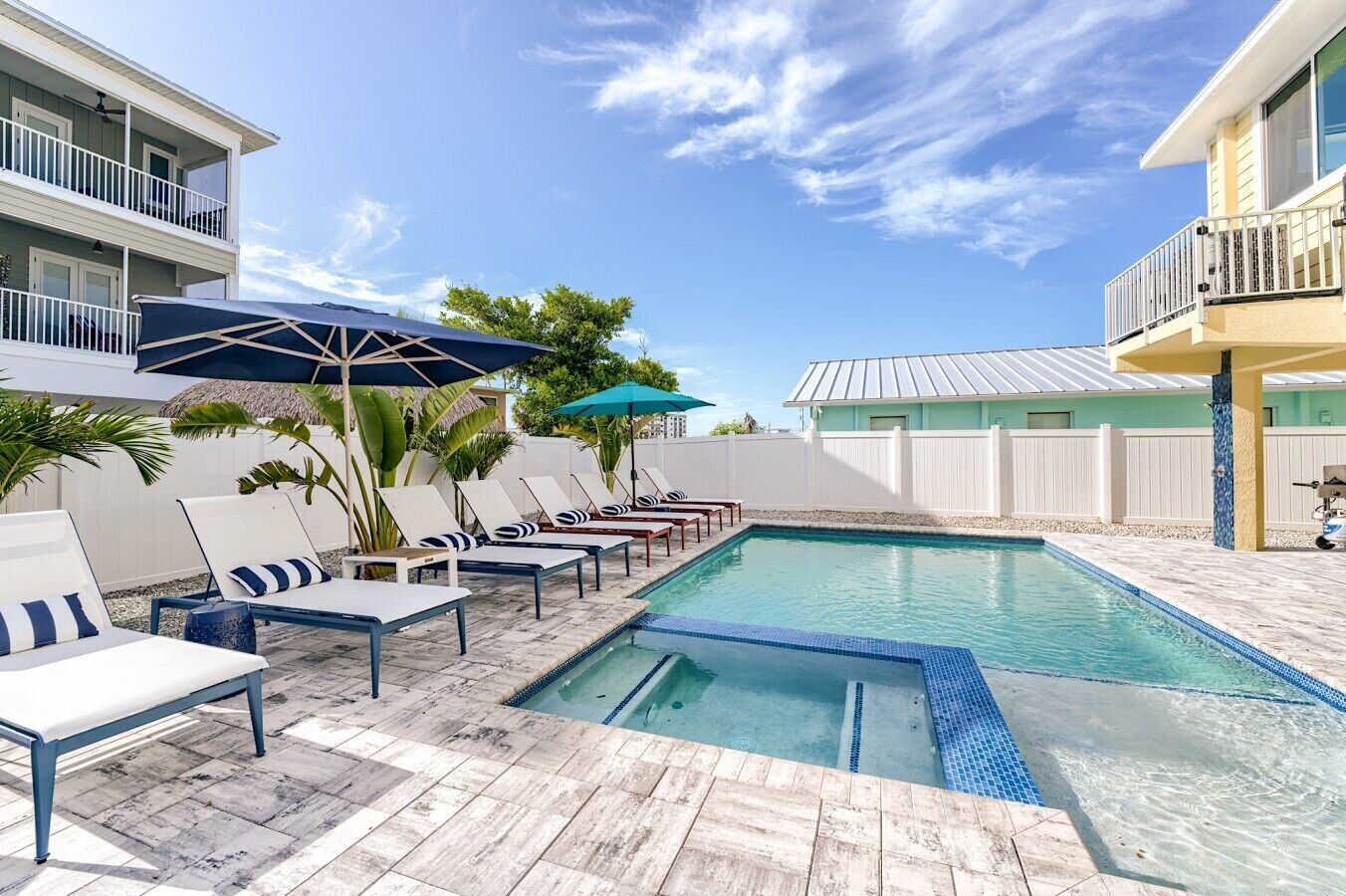 A swimming pool with lounge chairs and umbrellas, surrounded by a white fence and buildings.