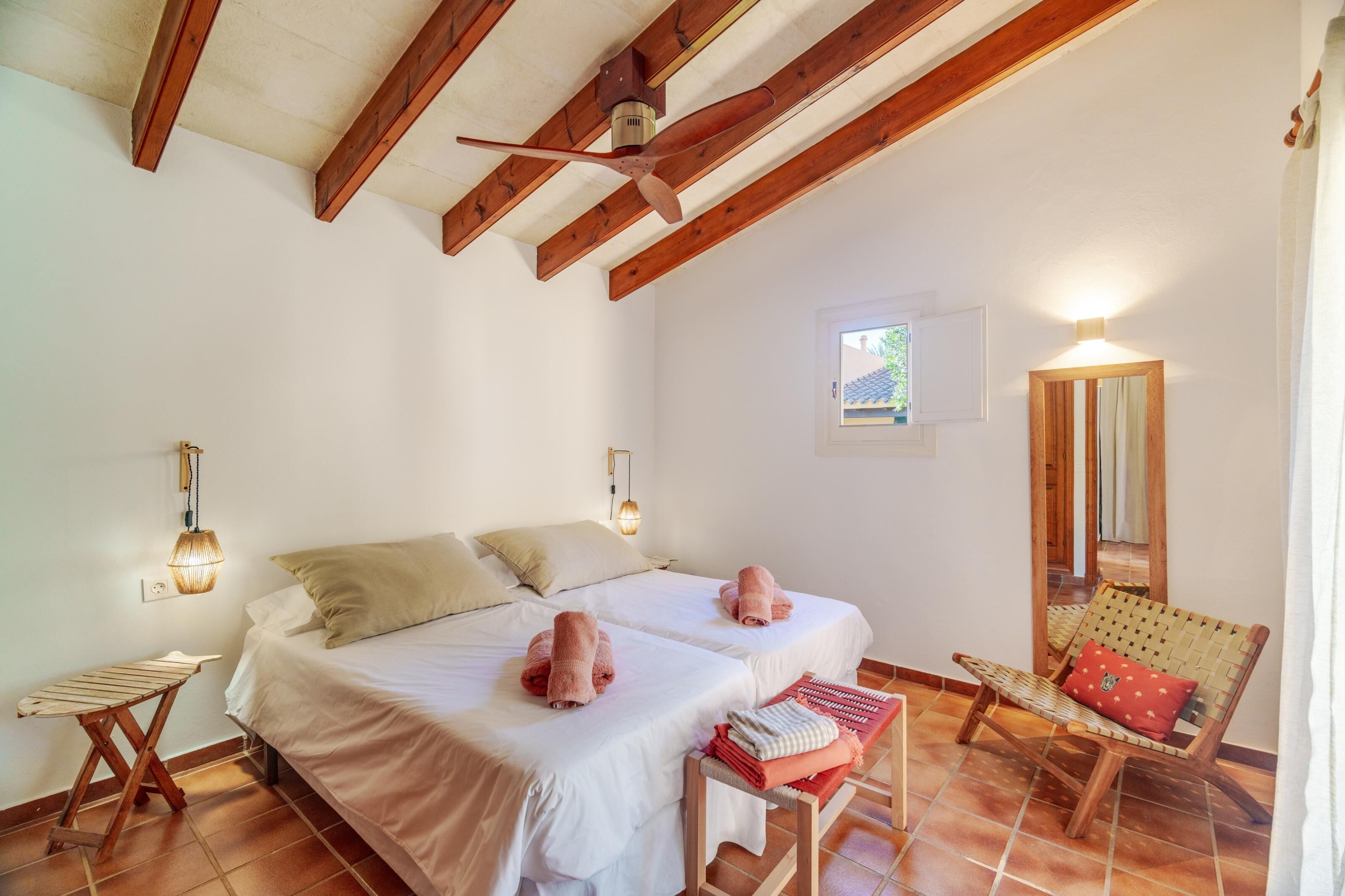 Ein Schlafzimmer mit zwei Betten, Holzboden, einem kleinen Fenster, einem wandmontierten Spiegel und einer Holzbank mit Kissen.