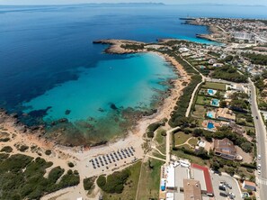 Vista aérea de una zona costera con playa, aguas azules cristalinas y un área residencial con casas y una carretera.