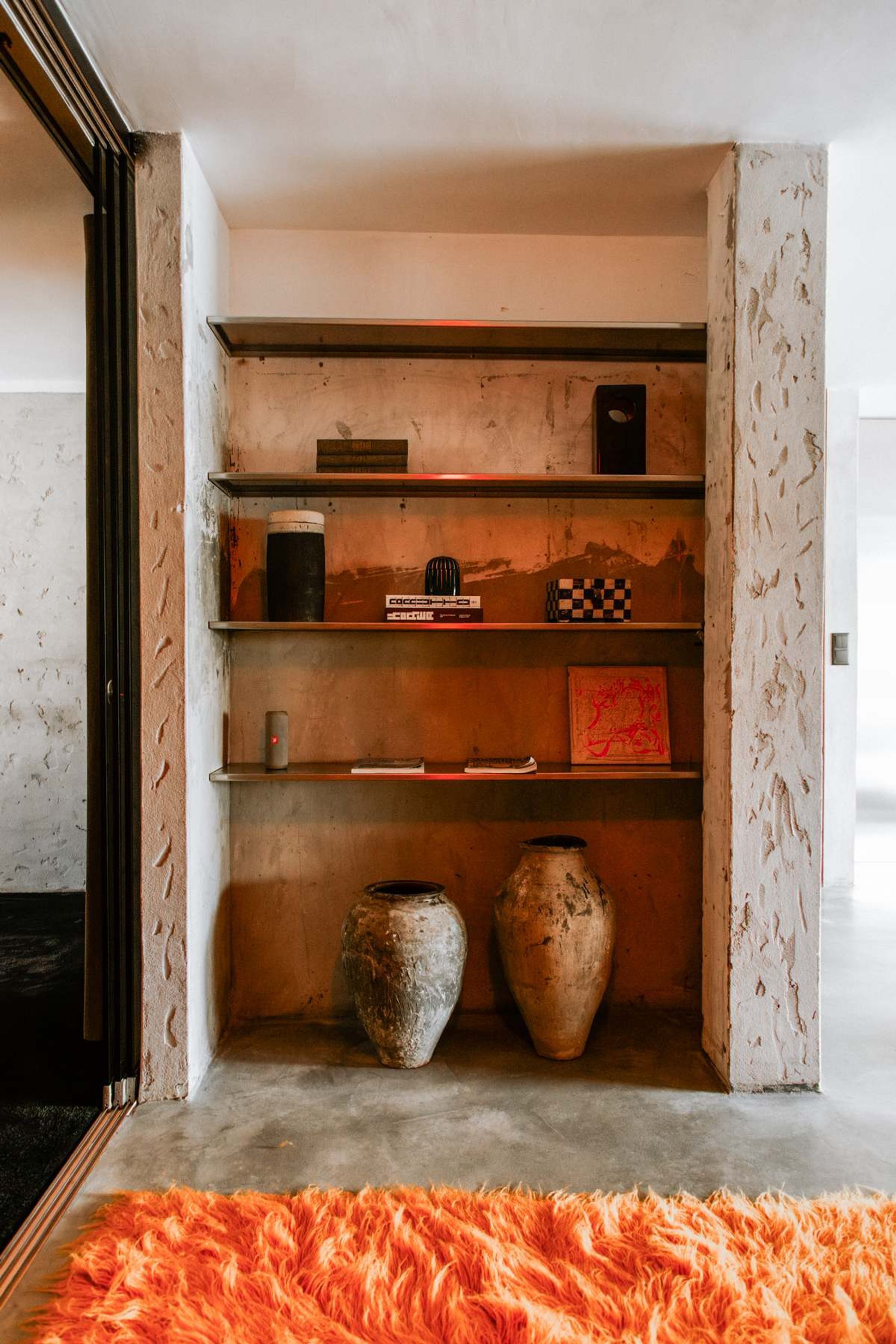 Una habitación con una pared de concreto, una repisa con varios objetos y una alfombra naranja grande.