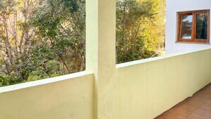 Ein Balkon mit gelber Wand und Fenster, mit Blick auf ein dichtes Grün der Bäume.