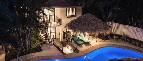 Ein zweistöckiges Haus mit Swimmingpool, einem überdachten Bereich mit Strohdach und einer Treppe, die zum oberen Stockwerk führt.