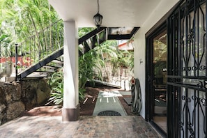 A covered outdoor area with a stone floor, a metal staircase, and a view of a garden with palm trees. - Queen Suite + Single Bed I Full Kitchen I Pool, Spa & Patio I Suite Three (Del Coco Beach)