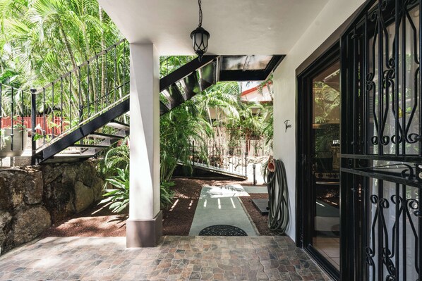 A covered outdoor area with a stone floor, a metal staircase, and a view of a garden with palm trees.