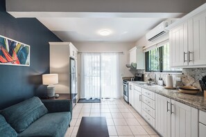 A modern kitchen with white cabinets, a marble countertop, and a dining area with a sofa and table. - Queen Suite + Single Bed I Full Kitchen I Pool, Spa & Patio I Suite Three (Del Coco Beach)