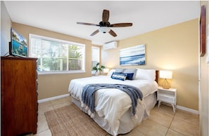 Une chambre à coucher avec un lit, un ventilateur de plafond, une table de chevet, une lampe et une fenêtre avec des stores.