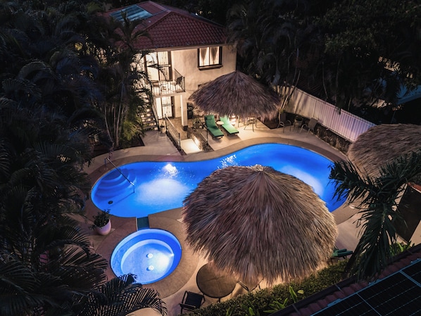 A swimming pool with a hot tub, surrounded by palm trees and a house with a red roof.