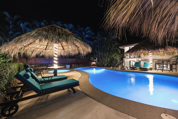 A swimming pool with lounge chairs, a thatched-roof structure, and a building with a similar roof.