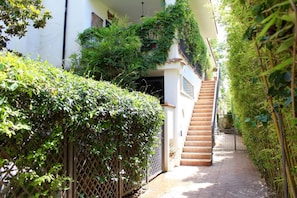 Ein schmaler Pfad, der zu einer Treppe führt, mit einem weißen Gebäude auf der einen Seite und dichtem Grünwerk auf der anderen.