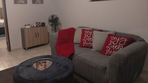 A living room with a grey sofa, red and white cushions, a small round ottoman, a wooden cabinet, and a fan.