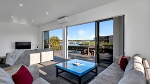 Un salon moderne avec une table basse en verre, des canapés gris, un téléviseur à écran plat et une vue sur une marina, visible à travers de grandes portes coulissantes.
