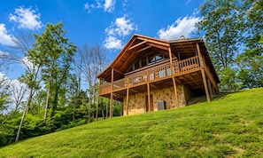 A wooden cabin with a balcony, surrounded by trees and a grassy hill. - Hard to Leave - Stunning Mountain Views, Pool Table, Games, Hot Tub! (Sevierville)