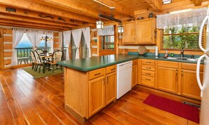 A wooden cabin interior with a kitchen area, wooden cabinets, a dining table with chairs, and a view through the windows. - Hard to Leave - Stunning Mountain Views, Pool Table, Games, Hot Tub! (Sevierville)