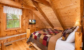 A wooden cabin bedroom with a bed, a bedside table, a TV, and a window with blinds. - Hard to Leave - Stunning Mountain Views, Pool Table, Games, Hot Tub! (Sevierville)