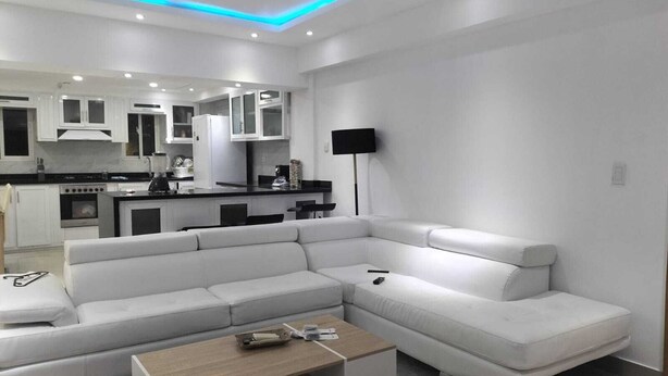 Ein modernes Wohnzimmer mit einem weißen Ecksofa, einem Holztisch und einer Küchenzeile mit weißen Schränken und einer schwarzen Arbeitsplatte.